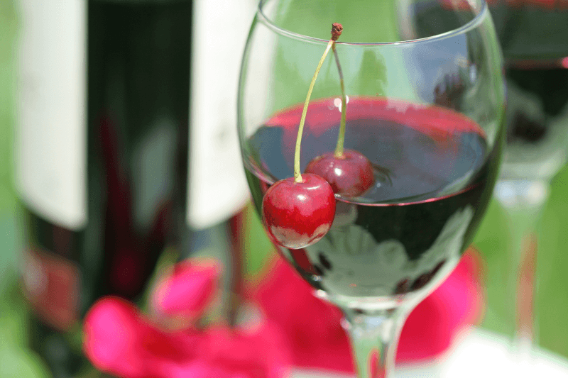 Glass of dark cherry wine garnished with fresh cherries beside wine bottles