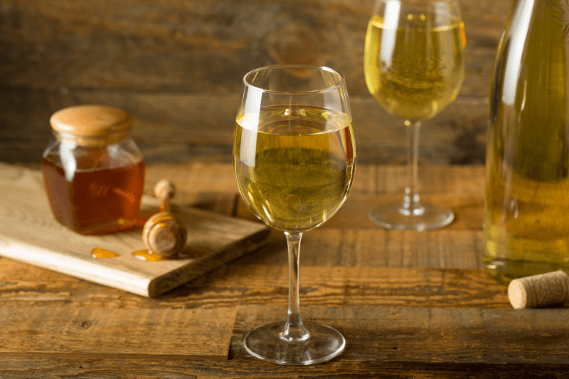 Glass of golden mead beside a honey jar and dipper on a rustic wooden table