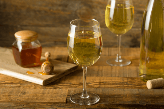 Glass of golden mead beside a honey jar and dipper on a rustic wooden table