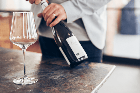 Person opening a bottle of sparkling wine beside an empty wine glass on a countertop