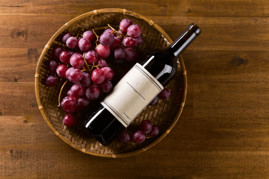 Red wine bottle placed in a basket with fresh grapes on a wooden table
