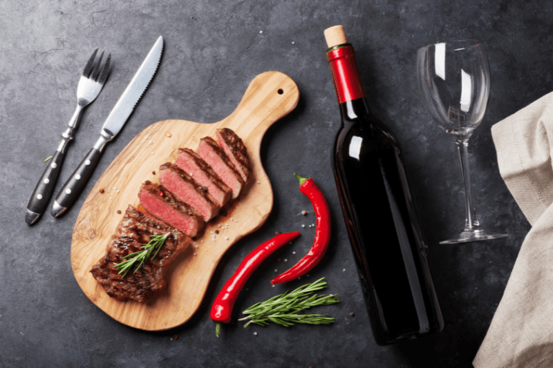 Sliced steak on a wooden board with red wine, herbs, chili peppers, and an empty wine glass